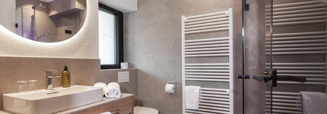 A modern bathroom with an elegant shower and a sink. The walls are bright and minimally designed, featuring a towel warmer and various toiletries. - Hotel Hubertus - Sölden