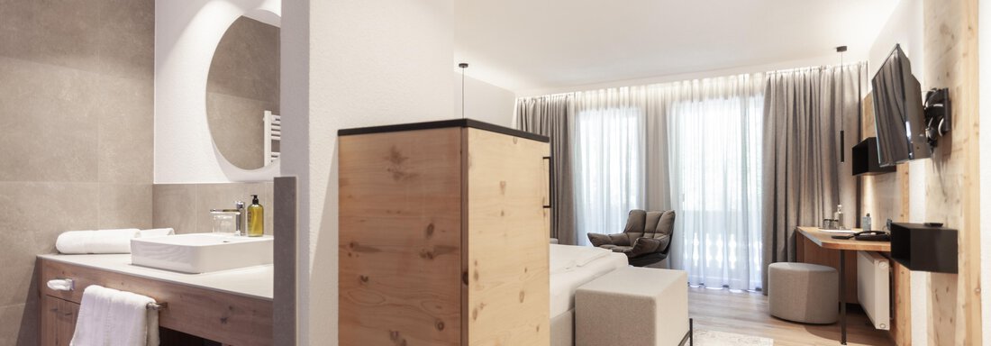 A modern hotel room with an adjoining bathroom. The space is bright and stylishly furnished with wooden and neutral colors. - Hotel Hubertus - Sölden