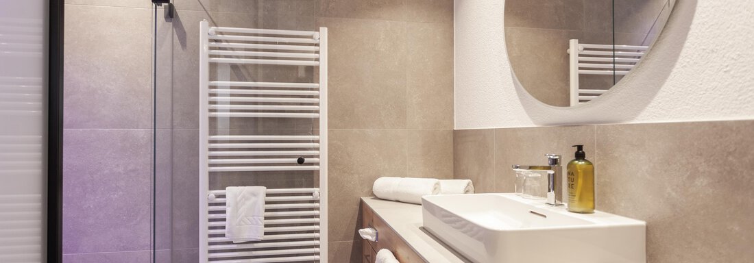 A modern bathroom with an elegant shower cabin and a round mirror. The walls are in neutral colors and there is a simple sink unit. - Hotel Hubertus - Sölden