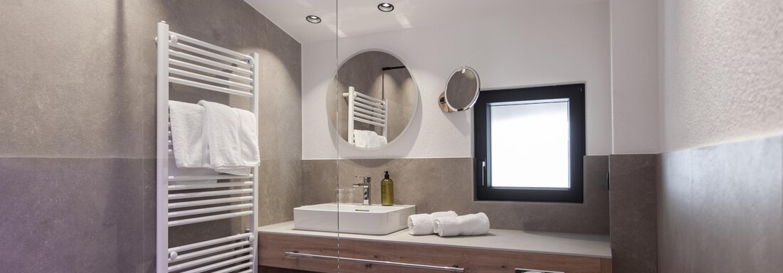 A modern bathroom with gray tiles and a wooden wash basin. There is a large mirror and a towel rack. - Hotel Hubertus - Sölden