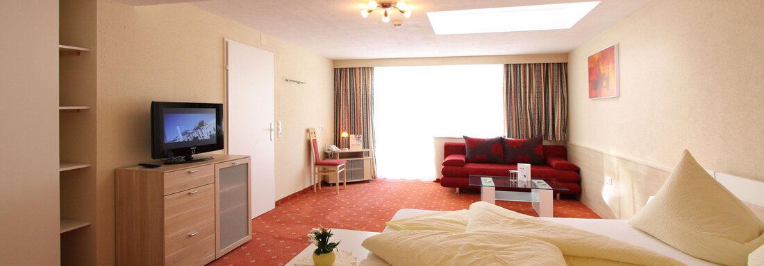 Ein modernes Zimmer mit einem Doppelbett, einem TV und einem gemütlichen roten Sofa. Große Fenster sorgen für viel Tageslicht. - Hotelappart Peter - Sölden