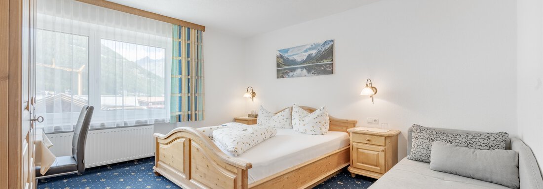 Doppelzimmer | © Stefanie Lohmann, Pension Charly (CC C) A cozy bedroom with a double bed and a gray sofa. The room has large windows and a warm wood decor. - Pension Charly - Sölden
