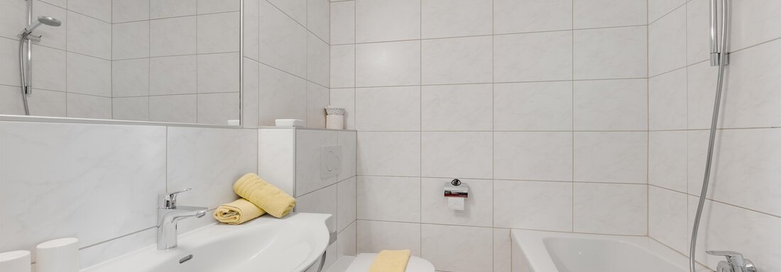 A modern bathroom with white tiles and a bathtub. There is also a sink and fresh towels in yellow. - Pension Charly - Sölden