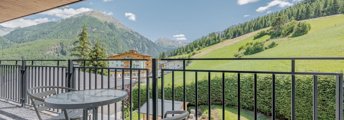 A balcony with a table and chairs offers a view of green hills and mountains. The sky is clear and sunny, ideal for relaxed moments outdoors. - Pension Charly - Sölden