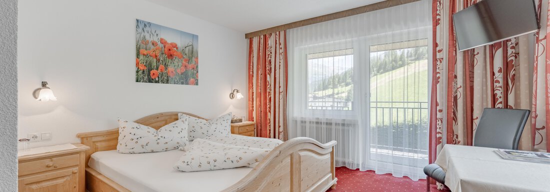 A cozy room with a double bed and colorful curtains. There is a desk and a balcony with a view outside. - Pension Charly - Sölden