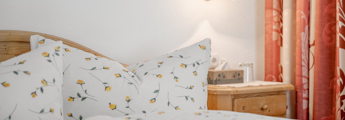 A cozy bedroom with a wooden bed and pretty pillows. The room is brightly decorated and has a classic nightstand with a lamp. - Pension Charly - Sölden