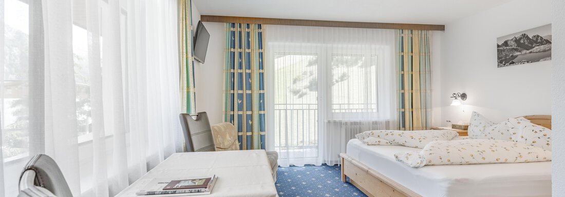 A bright room with two beds and a table.  
Large windows overlooking a balcony provide plenty of daylight. - Pension Charly - Sölden