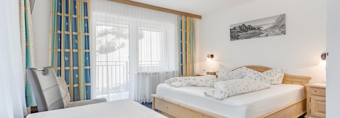 A cozy bedroom with a large bed and light curtains.  
The room has a table and a balcony with plenty of natural light. - Pension Charly - Sölden
