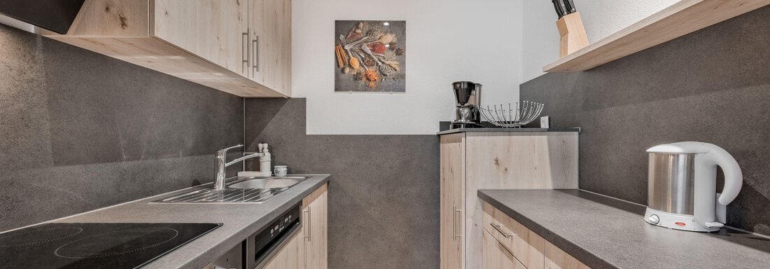 A modern kitchen with wooden cabinets and a dark countertop. It is well-equipped with appliances such as a stove, a kettle, and a coffee maker. - Pension Charly - Sölden