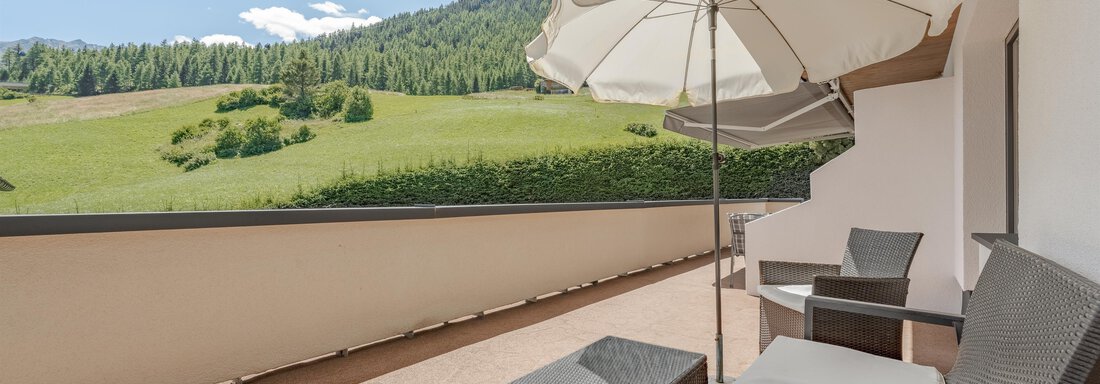 A cozy terrace with modern furniture and a sun umbrella. In the background, green meadows and mountains can be seen under a blue sky. - Pension Charly - Sölden