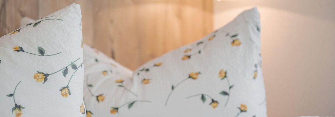 A cozy bed with pillows and bed linen that features delicate floral patterns. In the background, a warm wall lamp can be seen. - Pension Charly - Sölden