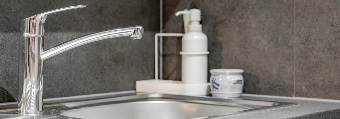 A modern kitchen sink with an elegant faucet. On the countertop next to the sink are a soap dispenser and a small container. - Pension Charly - Sölden