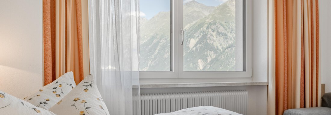 A cozy bedroom with a window through which the mountains are visible. The bedding has a floral pattern and harmonizes with the orange curtains. - Pension Charly - Sölden