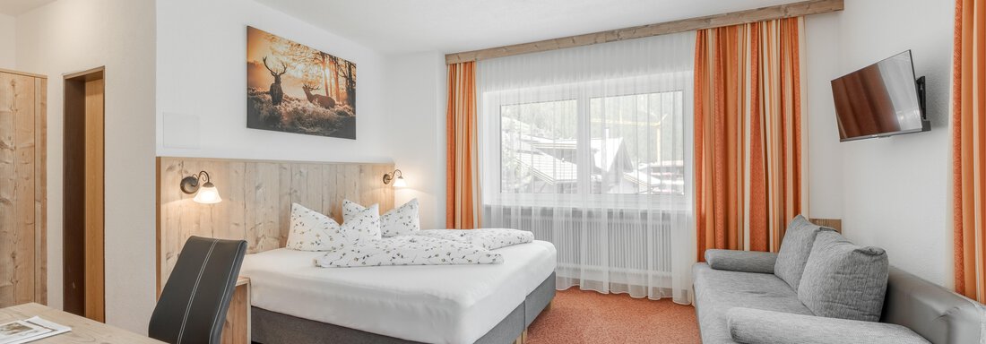 A bright and modern room with a double bed and large window. There is a desk and a couch, as well as orange curtains that create a cozy atmosphere. - Pension Charly - Sölden