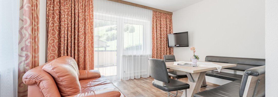 A modern living room with an orange sofa and a table. Large windows let in plenty of light and provide access to a balcony. - Pension Charly - Sölden