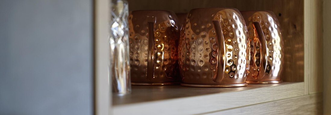 A row of copper-colored cups is standing on a wooden shelf. Next to it lies a transparent glass. - Pension "Zur alten Mühle" - Sölden