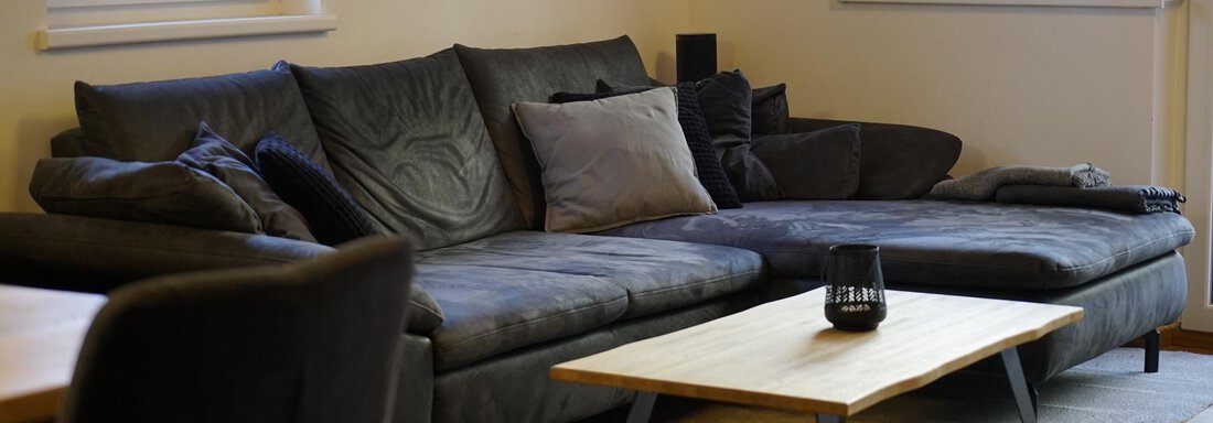 A modern living room with a comfortable, dark gray sofa and a simple wooden table. The walls are light, and there are large windows that provide plenty of natural light. - Pension "Zur alten Mühle" - Sölden