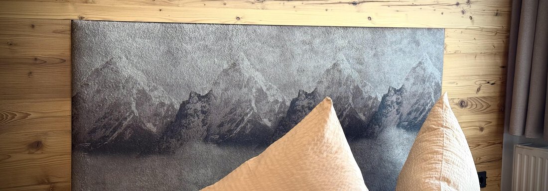 Ein modernes Schlafzimmer mit einer Holzverkleidung und einer Kopfteilgestaltung, die Berge darstellt. Die zwei großen Kissen fügen eine gemütliche Note hinzu. - Pension "Zur alten Mühle" - Sölden