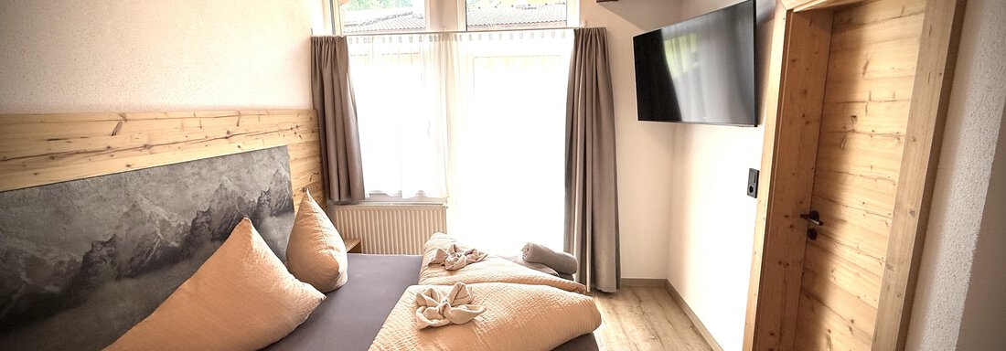 Ein modernes Schlafzimmer mit Holzdekor und großen Fenstern. Es gibt ein bequemes Bett mit großen Kissen und einen Fernseher an der Wand. - Pension "Zur alten Mühle" - Sölden