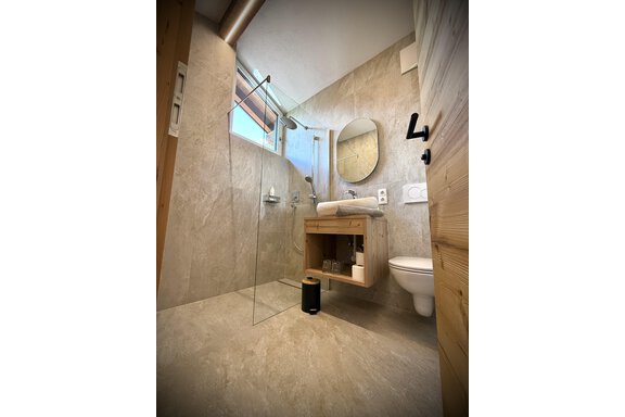 A modern bathroom with a bright stone floor. It features a glass shower partition, a round mirror, and a wooden washbasin. - Pension "Zur alten Mühle" - Sölden
