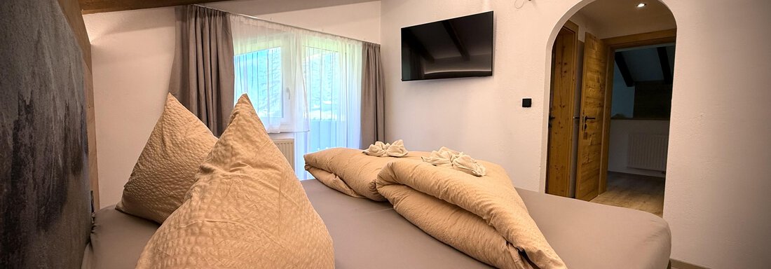 Ein gemütliches Schlafzimmer mit einem großen Bett und vielen Kissen. Es gibt ein Fenster mit Vorhängen und einen Fernseher an der Wand. - Pension "Zur alten Mühle" - Sölden