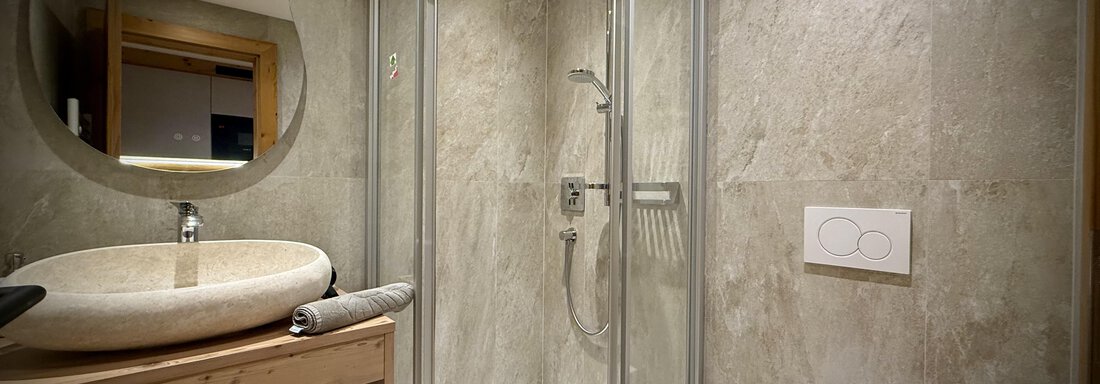 A modern bathroom with a glass shower and a round mirror. The sink is made of stone, and the walls are kept in neutral tones. - Pension "Zur alten Mühle" - Sölden