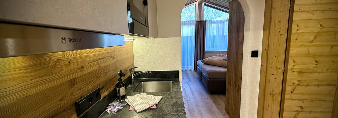 A modern kitchen with wooden details and a dark workspace. In the background, a cozy bedroom is visible. - Pension "Zur alten Mühle" - Sölden