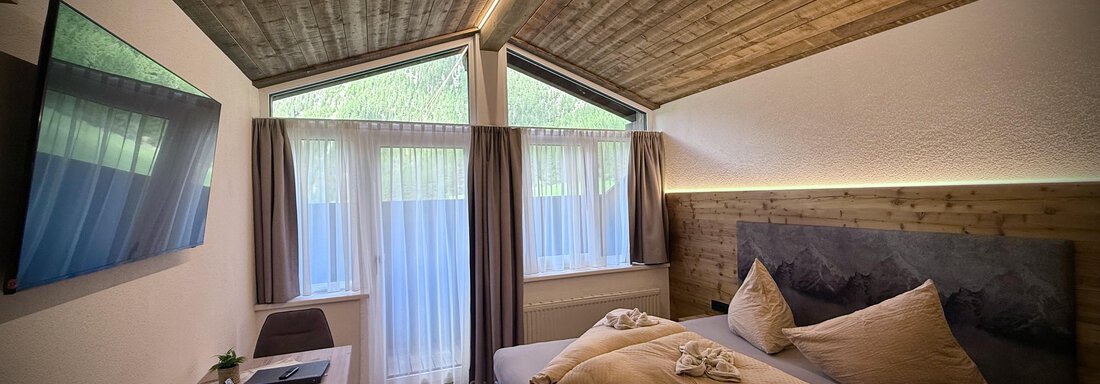 A modern room with wooden ceiling and large windows. It is bright and comfortably furnished with a double bed and a desk. - Pension "Zur alten Mühle" - Sölden