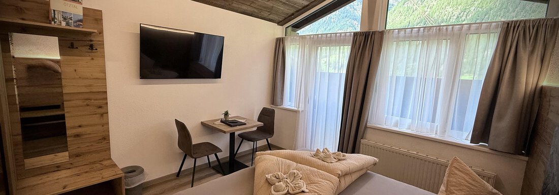 A modern room with a wooden ceiling and large windows.  
It is minimally furnished with a double bed and a table with chairs. - Pension "Zur alten Mühle" - Sölden