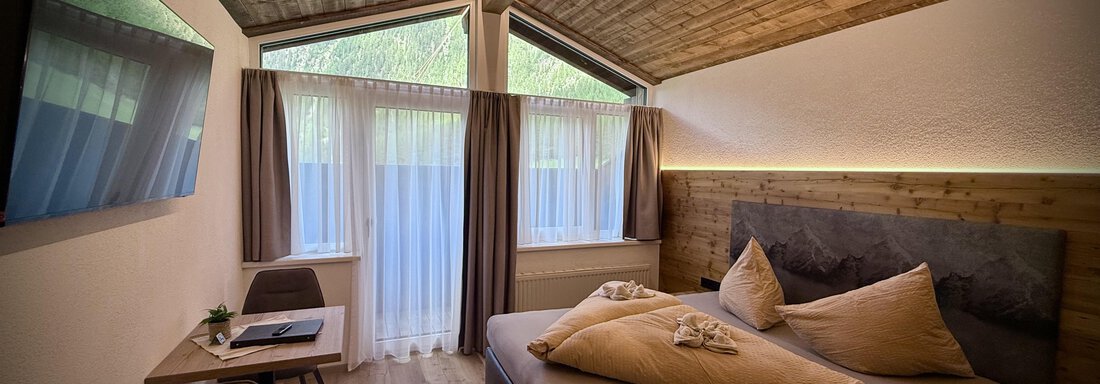Ein modernes Schlafzimmer mit Holzdecke und großen Fenstern. Es ist hell und gemütlich mit einem großen Bett und einem kleinen Arbeitsplatz. - Pension "Zur alten Mühle" - Sölden
