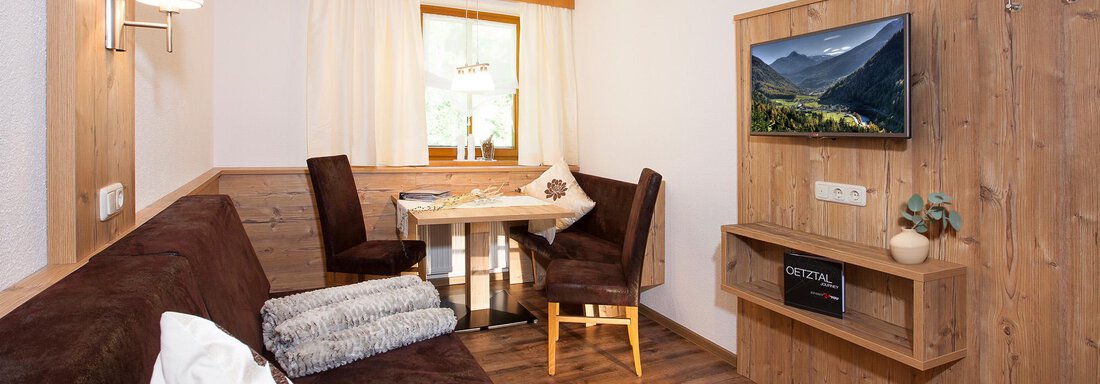 A cozy living space with a sofa, a table, and two chairs. The room is bright and friendly, with a television on the wall and warm wooden details. - Trofana Appartements - Sölden