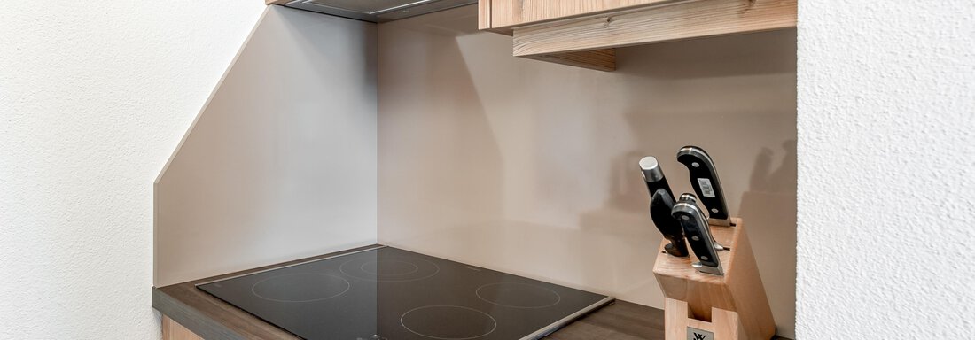 Eine moderne Küchenzeile mit Holzfronten und einem Keramikkochfeld. Auf der Arbeitsfläche steht ein Messerblock mit mehreren Messern. - Villa Castelbell - Sölden