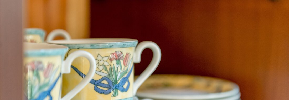 Gestapelte Tassen mit buntem Blumendesign und einige Teller im Hintergrund. Die Tassen stehen in einem Holzregal. - Villa Castelbell - Sölden