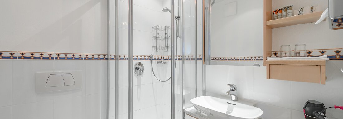 Ein modernes Badezimmer mit einer transparenten Duschnische und einem Waschtisch. Die Wände sind weiß gefliest und es gibt Holzregale für Pflegeprodukte. - Villa Castelbell - Sölden