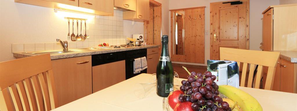 Eine moderne Küche mit Holzregalen und einem Esstisch. Auf dem Tisch steht eine Obstschale mit Trauben, Äpfeln und Bananen sowie eine Flasche Sekt. - Waldele - Sölden