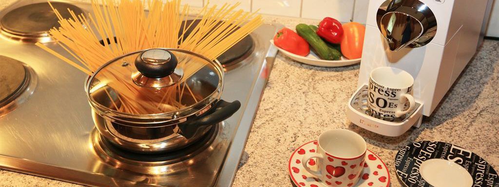 Ein Kochtopf mit Spaghetti auf einem Herd steht neben einer Kaffeemaschine. Auf dem Tisch sind zwei Tassen, eine Schale mit Gemüse und eine Untertasse dekoriert. - Waldele - Sölden