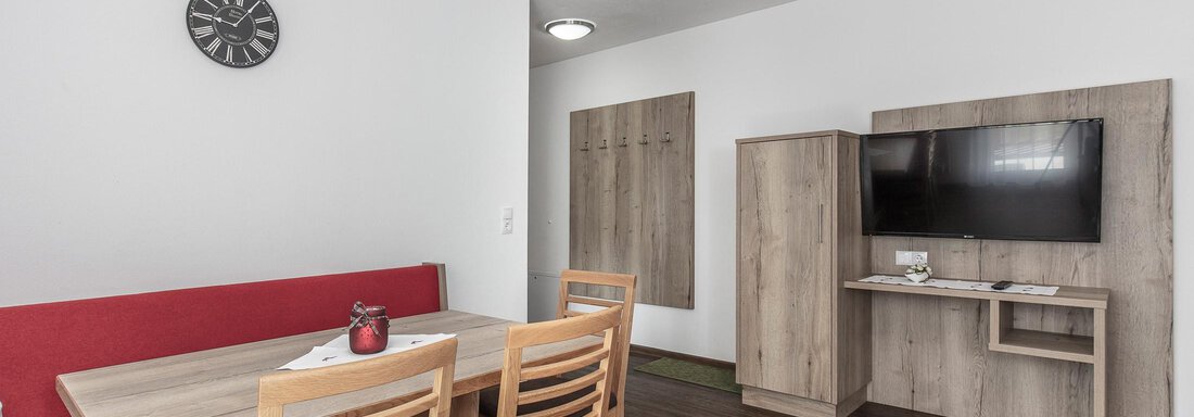 Ein modernes Wohnzimmer mit einem Esstisch, Stühlen und einem Fernseher. Die Wände sind hell, und es gibt eine rote Couch sowie eine Wanduhr. - Zenz´n Hof - Sölden