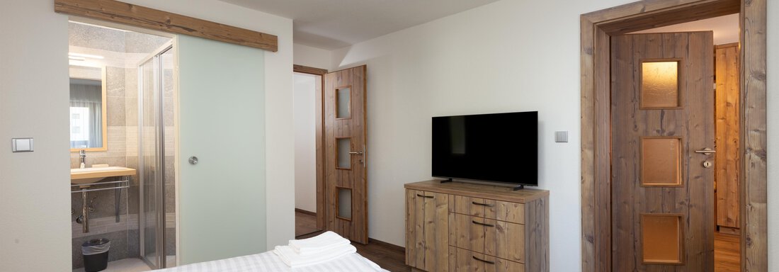 A modern bedroom with a double bed and a wooden furniture dresser. In the background, there is a bathroom with a glass wall and a closed door. - Garni Stefani - Vent