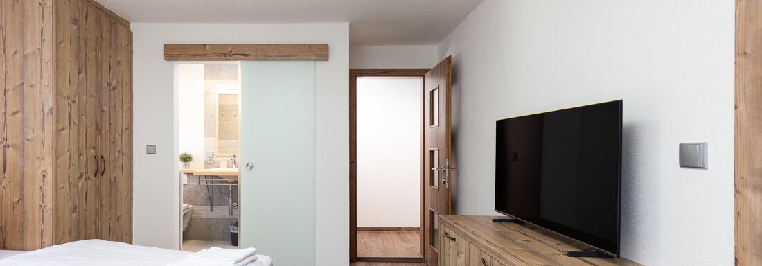 A modern bedroom with wooden paneling and a large television. The walls are light-colored, and there is access to the bathroom. - Garni Stefani - Vent