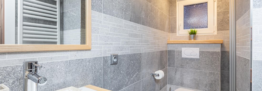A modern bathroom with grey tiles and a wooden washbasin. A small window frame with a plant adds a fresh accent. - Garni Stefani - Vent