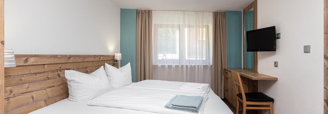 A modern bedroom with a wooden double bed and a desk. The room is bright and friendly with a window and a television on the wall. - Garni Stefani - Vent