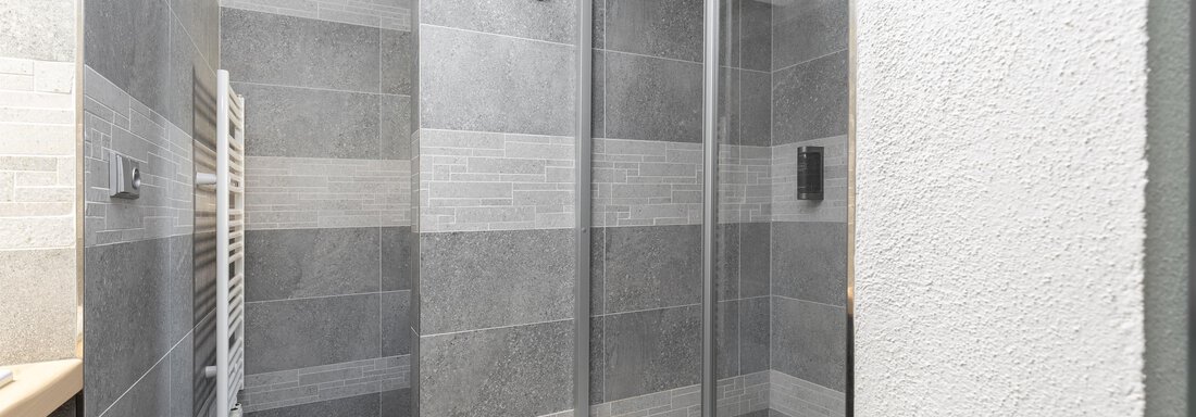 A modern bathroom with a glass door shower. The walls are tiled in different shades of gray. - Garni Stefani - Vent