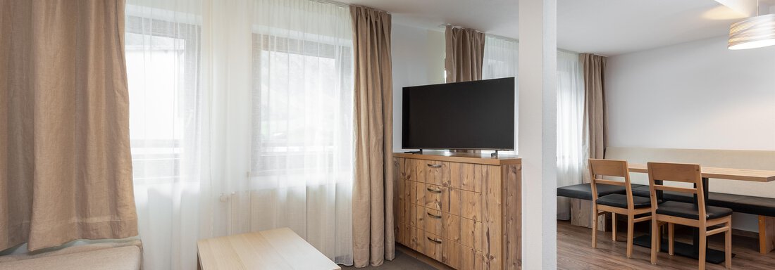 A modern living room with a cozy sofa and a television. A dining table with chairs is visible in the background. - Garni Stefani - Vent