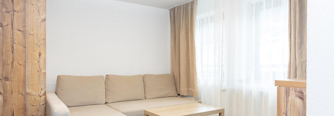 A cozy living room with a beige sofa and a wooden table. Large windows let in a lot of light and give the room a friendly atmosphere. - Garni Stefani - Vent
