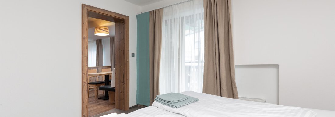 A simple and modern bedroom with a large window and bright light. The walls are painted in neutral colors, and there is a view of a dining area through the door. - Garni Stefani - Vent