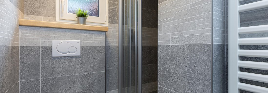 A modern bathroom with gray tiles and a shower. The small window provides daylight and a potted plant adds fresh accents. - Garni Stefani - Vent