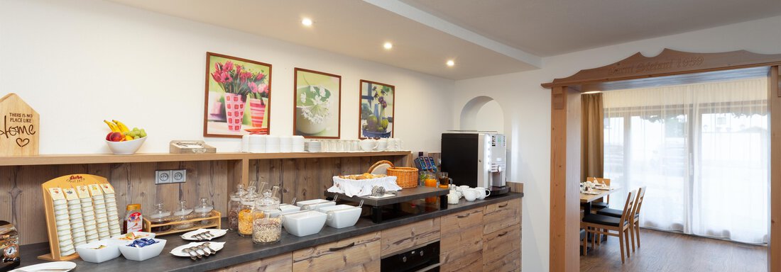 A modern kitchen with wooden cabinets and a buffet. On the table, there are many breakfast options and fresh fruit to be seen. - Garni Stefani - Vent