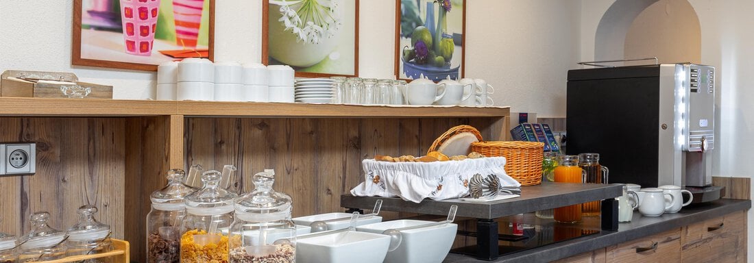 A breakfast buffet with various dishes in a wooden cabinet. In the background, there are pictures with floral motifs. - Garni Stefani - Vent