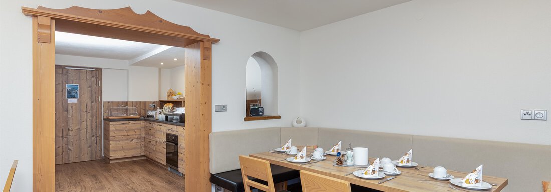 A cozy dining area with wooden chairs and a table. In the background, a kitchen can be seen. - Garni Stefani - Vent