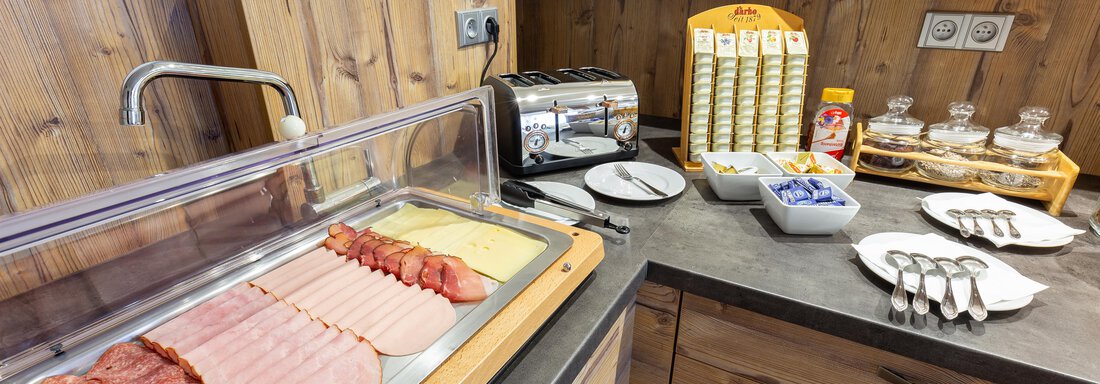 A breakfast table with a selection of cold cuts, toast, and jam. In the background, fruit and breakfast dishes can be seen. - Garni Stefani - Vent